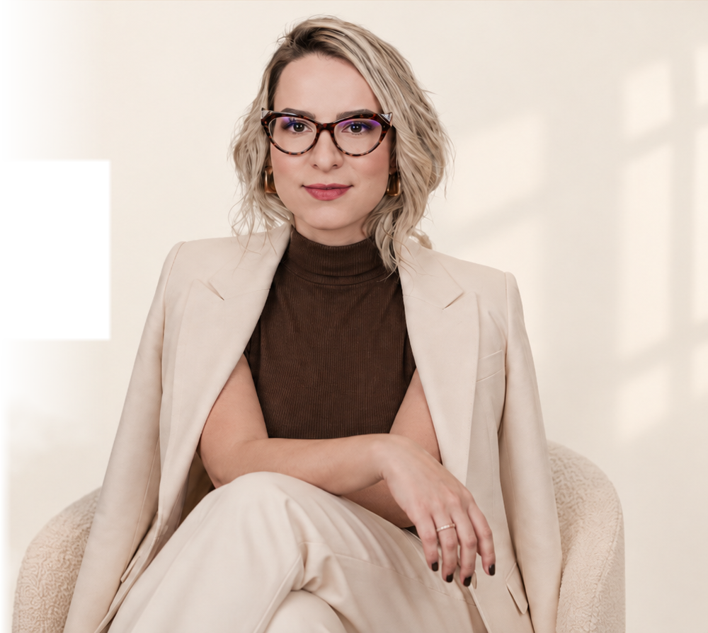 Laís Paschoal sentada em uma poltrona, de terno creme e óculos de acetato havana, olhando para a câmera
